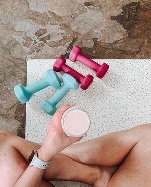 A pair of hands holding a container of a pinkish substance, with two dumbbells on a dotted mat. - Hotel Argento (St. Julian's)