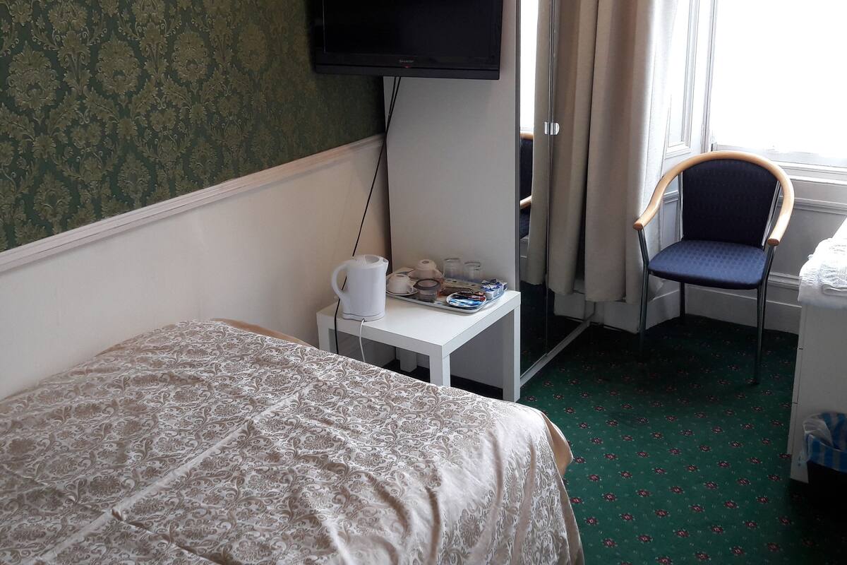 A hotel room with a bed, a small table with a kettle, a chair, a TV mounted on the wall, and a window with curtains.