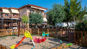 Ein Spielplatz mit Schaukeln und einer Rutsche, umgeben von einem Holzzaun und Bäumen.