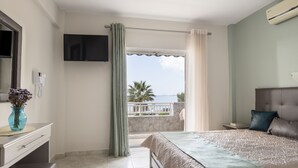 Ein Hotelzimmer mit Bett, einem an der Wand befestigten Fernseher, einem Badezimmer mit Toilette und einem Balkon mit Meerblick.