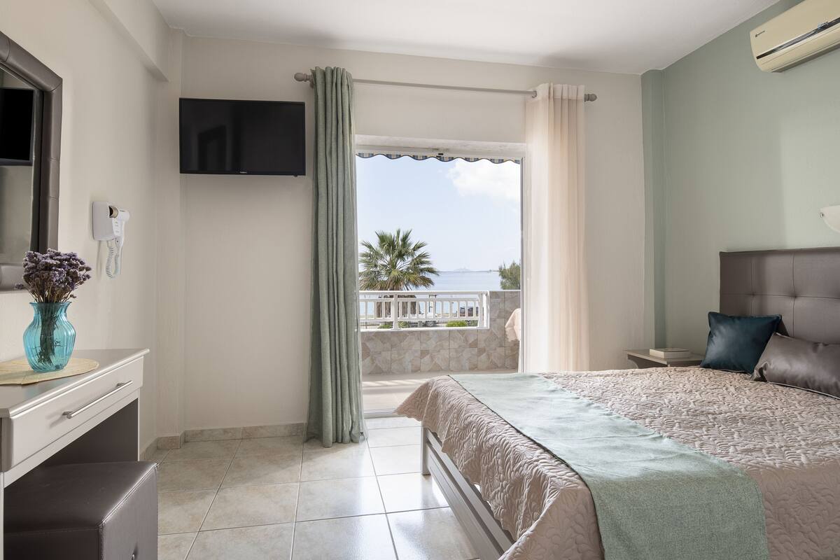 Habitación de hotel con cama, televisor montado en la pared, baño con inodoro y balcón con vista al mar.