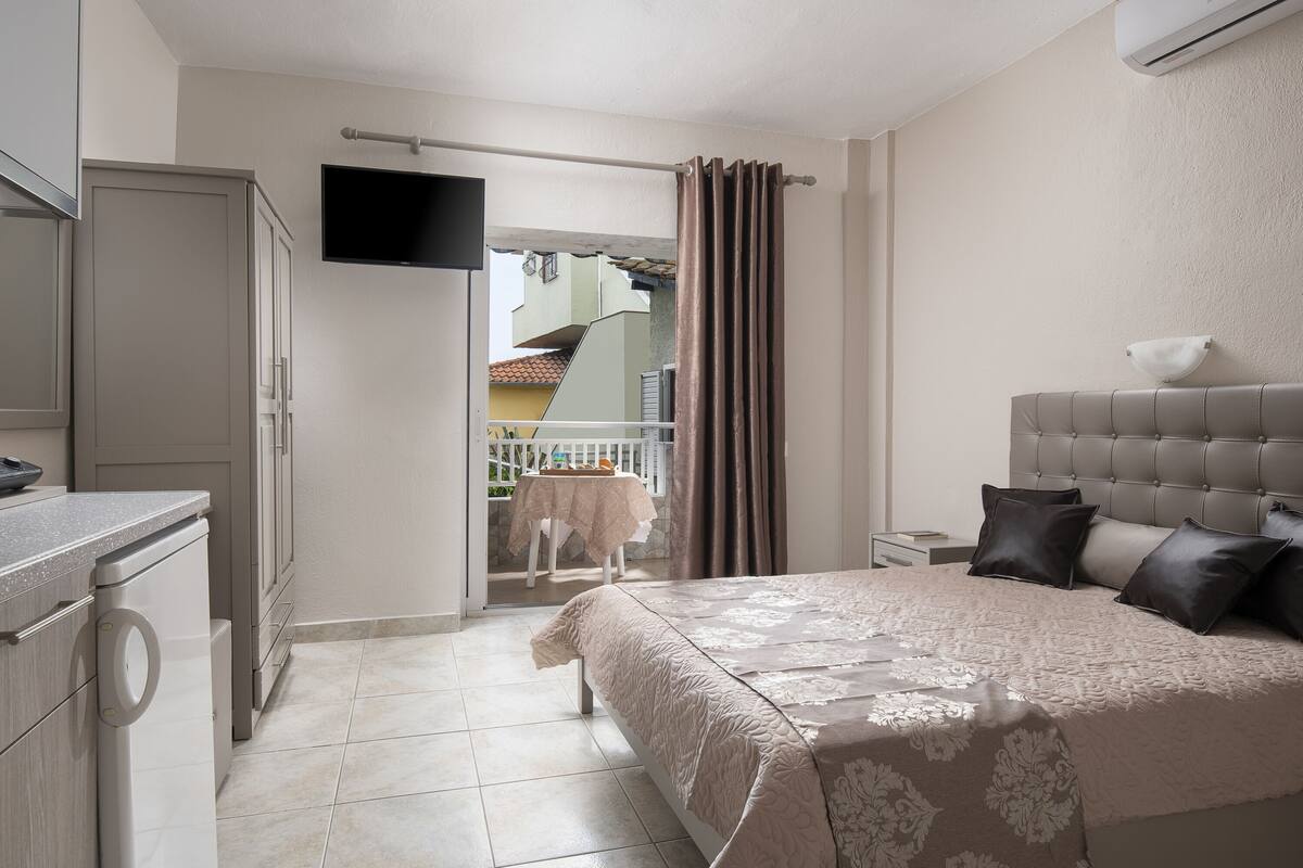 Un dormitorio con cama, televisión montada en la pared, lavadora y balcón con mesa y sillas.
