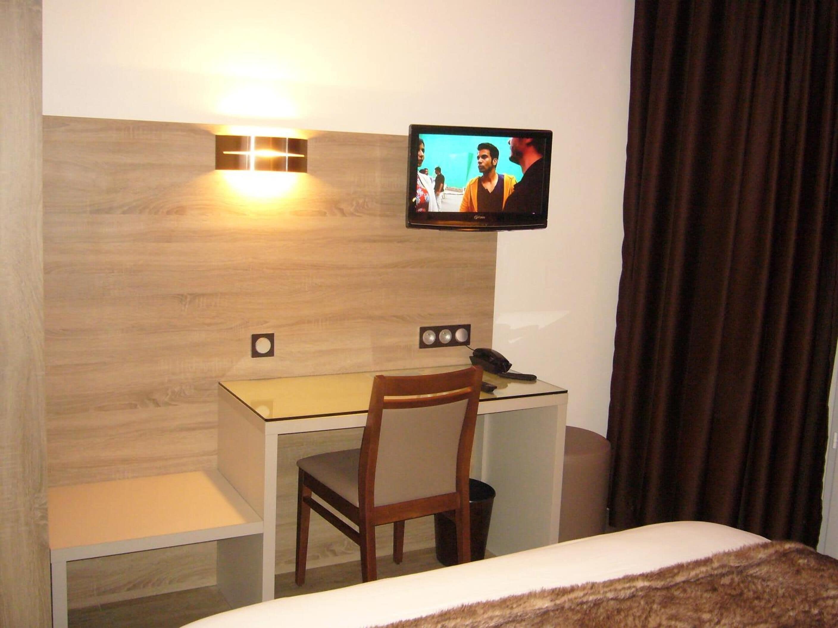 A hotel room with a wooden headboard, a desk with a chair, a bed, a television mounted on the wall, and a curtain.