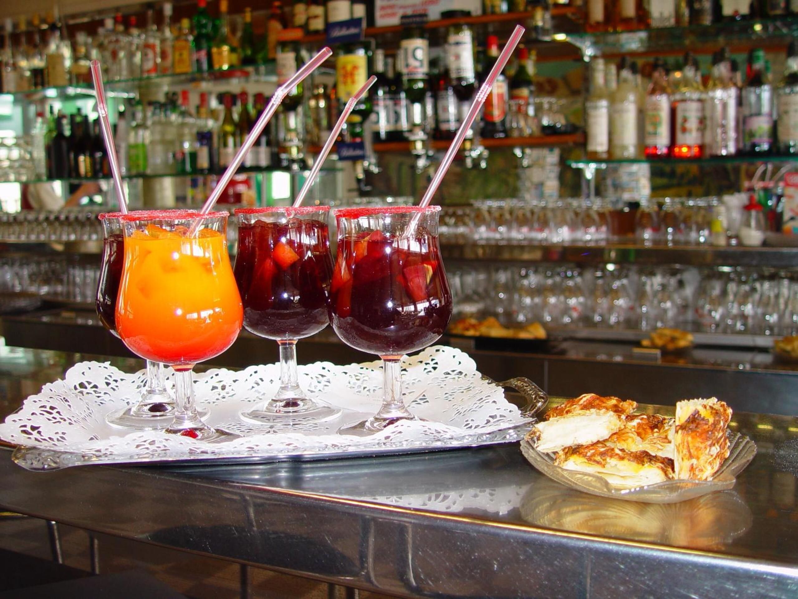 Una barra con tres bebidas en vasos, un plato de comida y un fondo de estanterías con botellas.