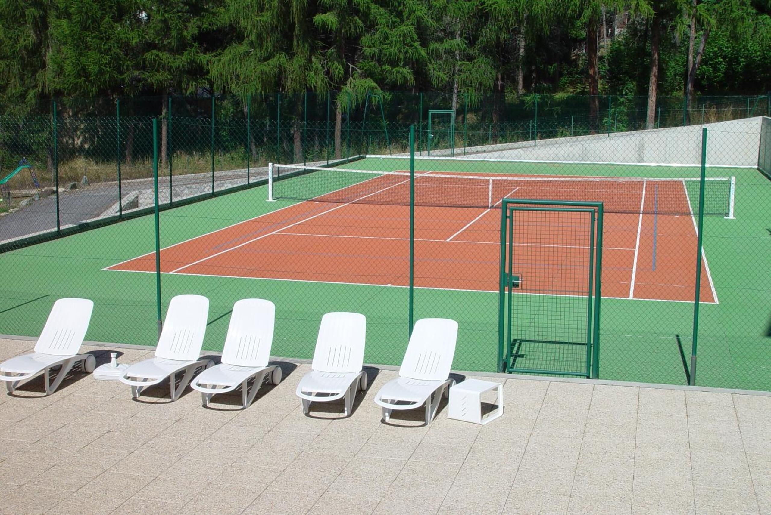 Una cancha de tenis con vallas verdes, tumbonas blancas y árboles al fondo.