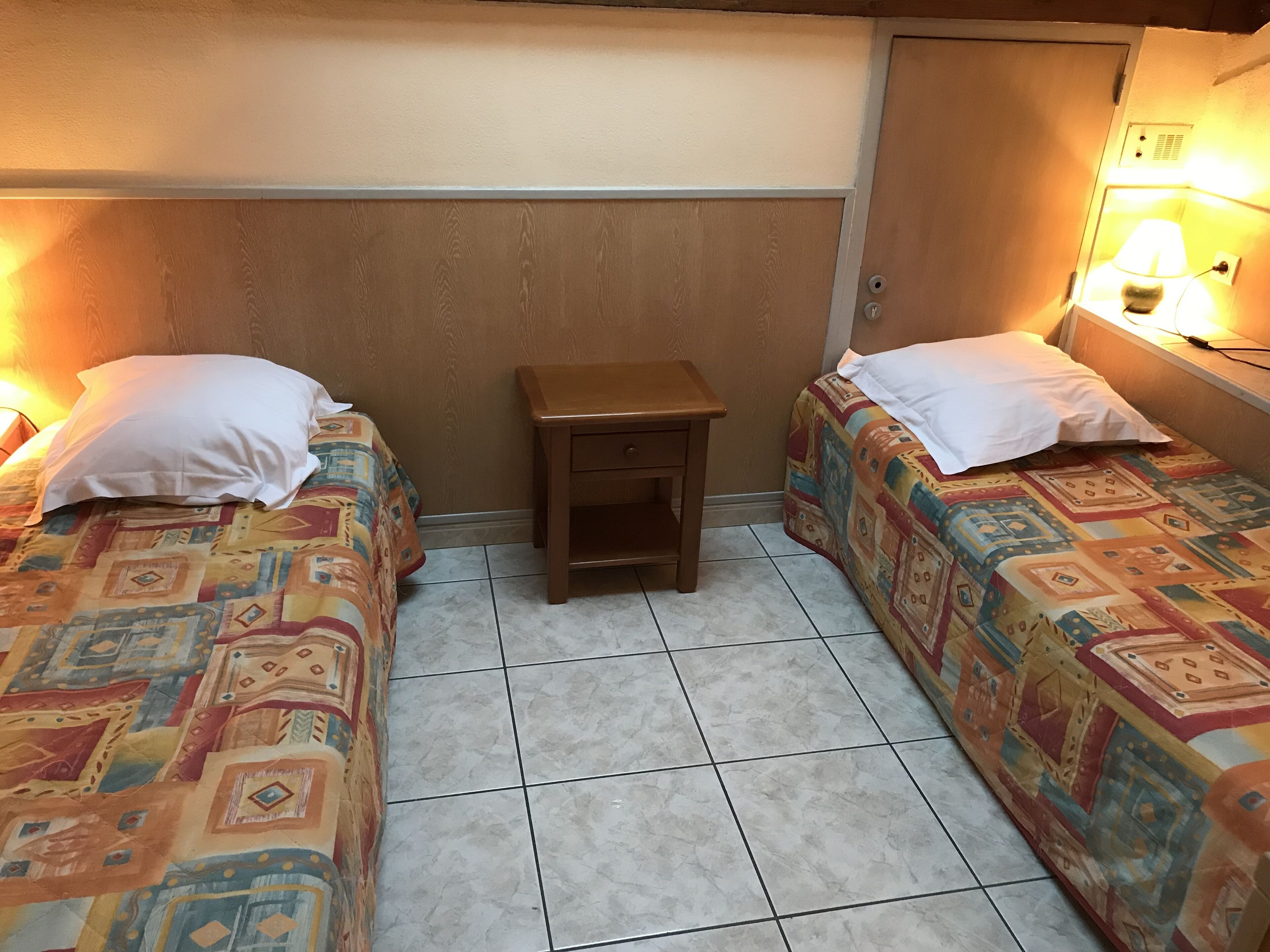A small, cozy room with two beds, each with patterned bedding and white pillows. There is a small wooden nightstand between the beds, and a wall-mounted lamp on the right side.