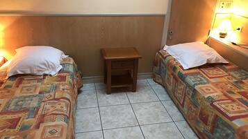 A small, cozy room with two beds, each with patterned bedding and white pillows. There is a small wooden nightstand between the beds, and a wall-mounted lamp on the right side.