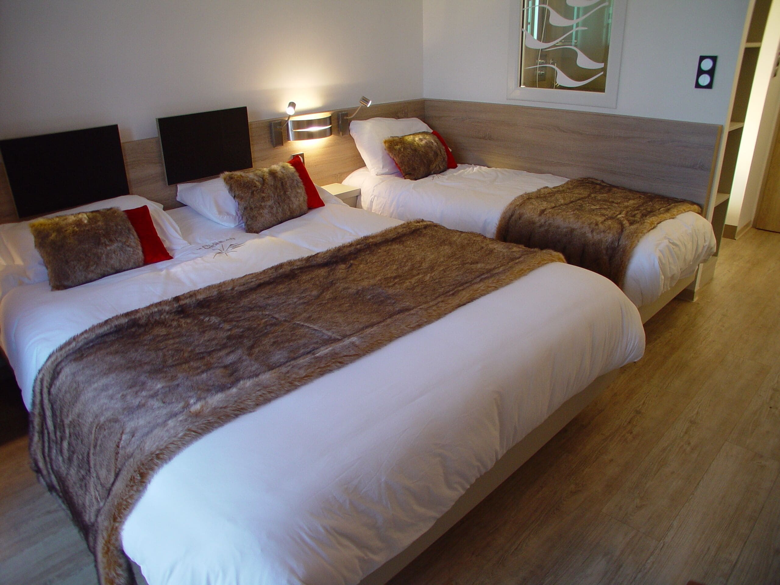 Habitación de hotel con dos camas, cada una con ropa blanca y mantas marrones. Hay un televisor montado en la pared y una mesita de noche con una lámpara.