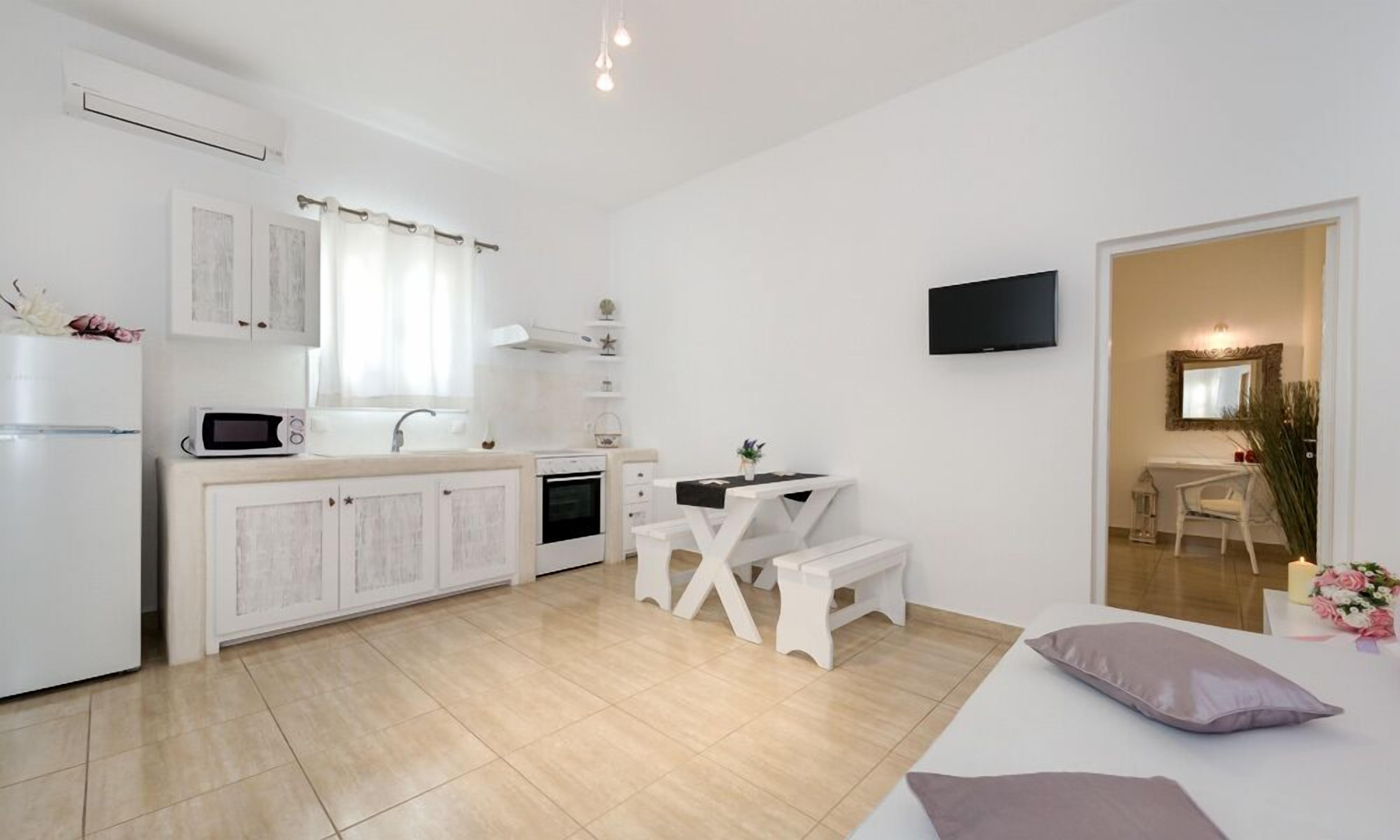 Una cocina moderna con armarios blancos, un microondas y un lavamanos. Hay una zona de comedor con una mesa y un banco blancos. Un televisor está montado en la pared, y se ve una cama con almohada al frente.