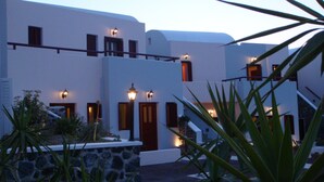 Un edificio blanco con varias puertas y ventanas, rodeado de follaje y muros de piedra.