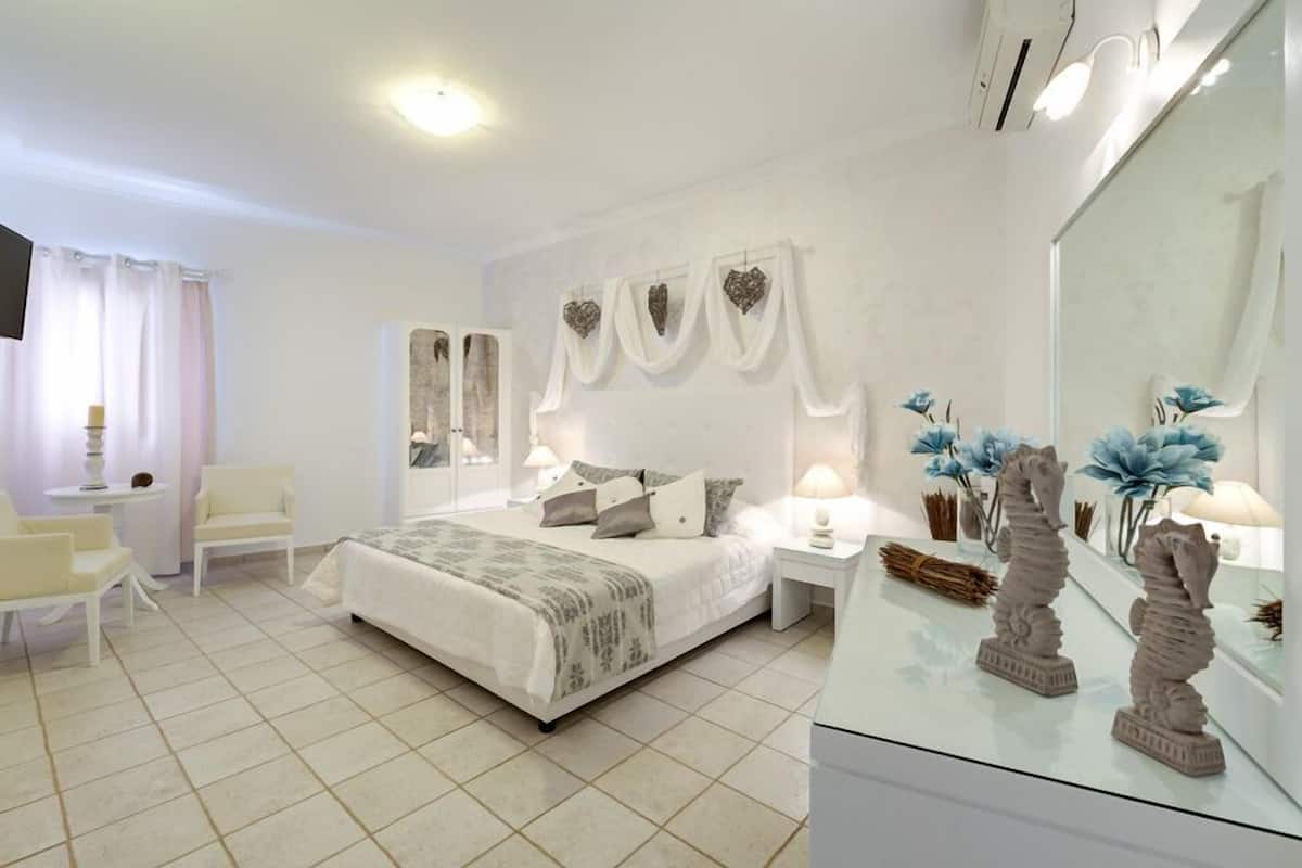 A modern bedroom with a large bed, a sitting area with a chair, a TV, and a vanity with decorative items.