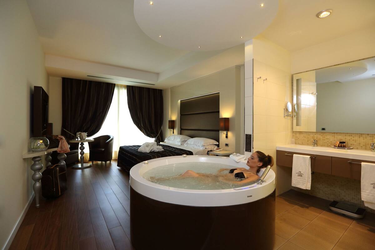 A modern hotel room with a large bathtub, a bed, a desk, and a bathroom with a large mirror and sink.