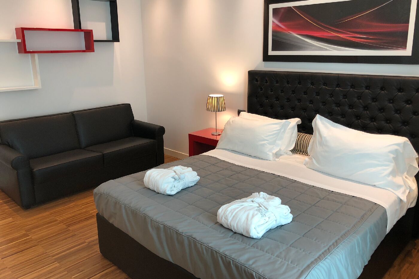 A hotel room with a large bed, a black sofa, a red nightstand, and a framed abstract painting on the wall.
