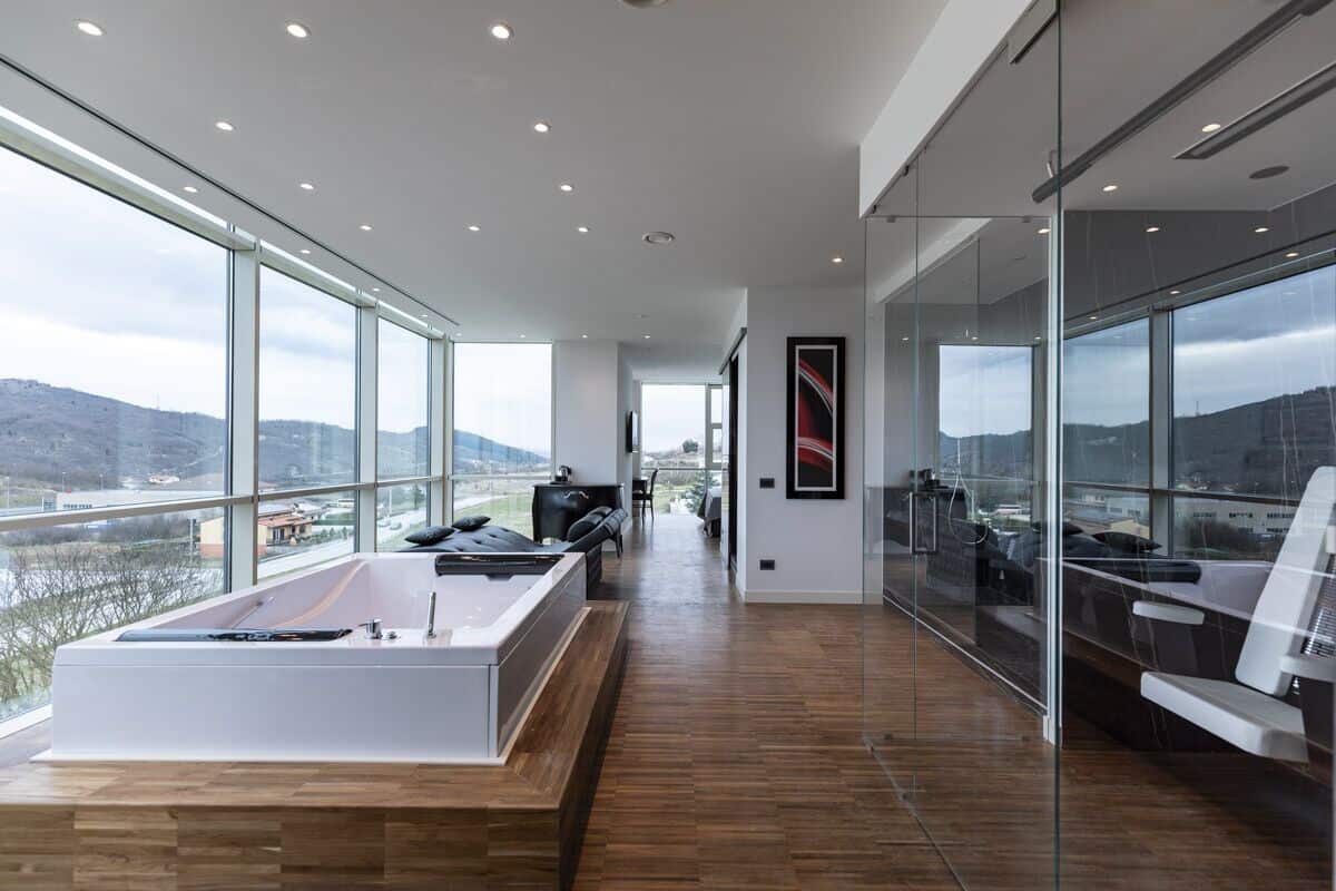 A modern hotel room with a large bathtub, a sofa, and a glass wall offering a city view.