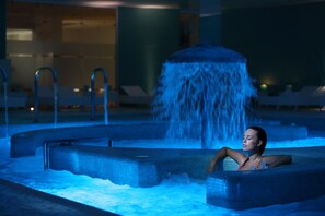 A person relaxing in a hot tub with a waterfall feature, surrounded by lounge chairs and a tiled pool area. - Fonte Del Benessere Resort Castelpetroso - Molise (Castelpetroso)