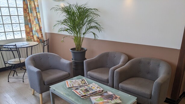 Una habitación con dos sillones grises, una mesa de centro de cristal con periódicos, una planta en maceta y una mesita redonda con una silla.