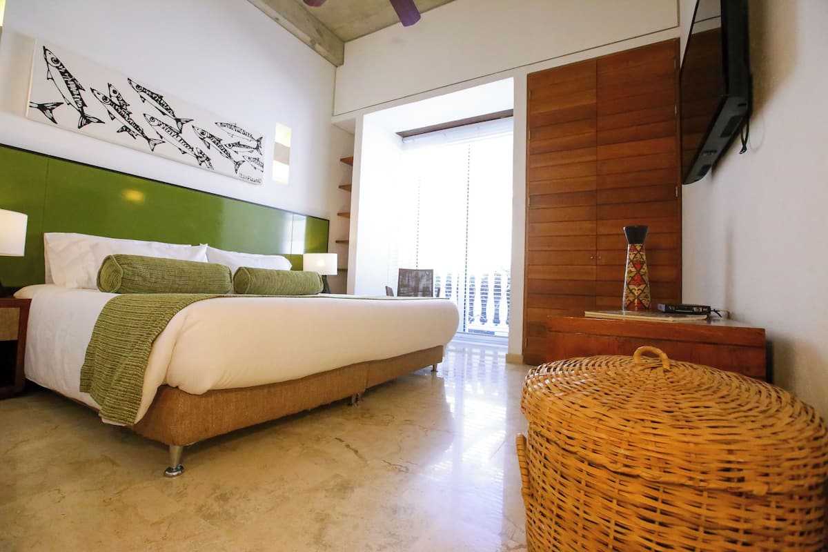 Un dormitorio con una cama, cabecera verde, televisor montado en la pared y una silla de mimbre.