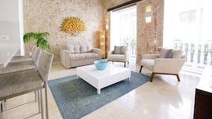 A modern living room with a white coffee table, two sofas, a dining area with a table and chairs, and a large window with blinds. - Casa Claver Loft Boutique Hotel (Cartagena)