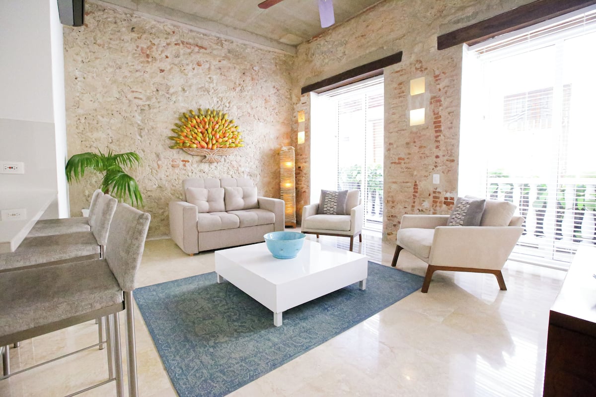 Una sala de estar moderna con una mesa de centro blanca, dos sofás, un comedor con una mesa y sillas, y un ventanal con persianas.