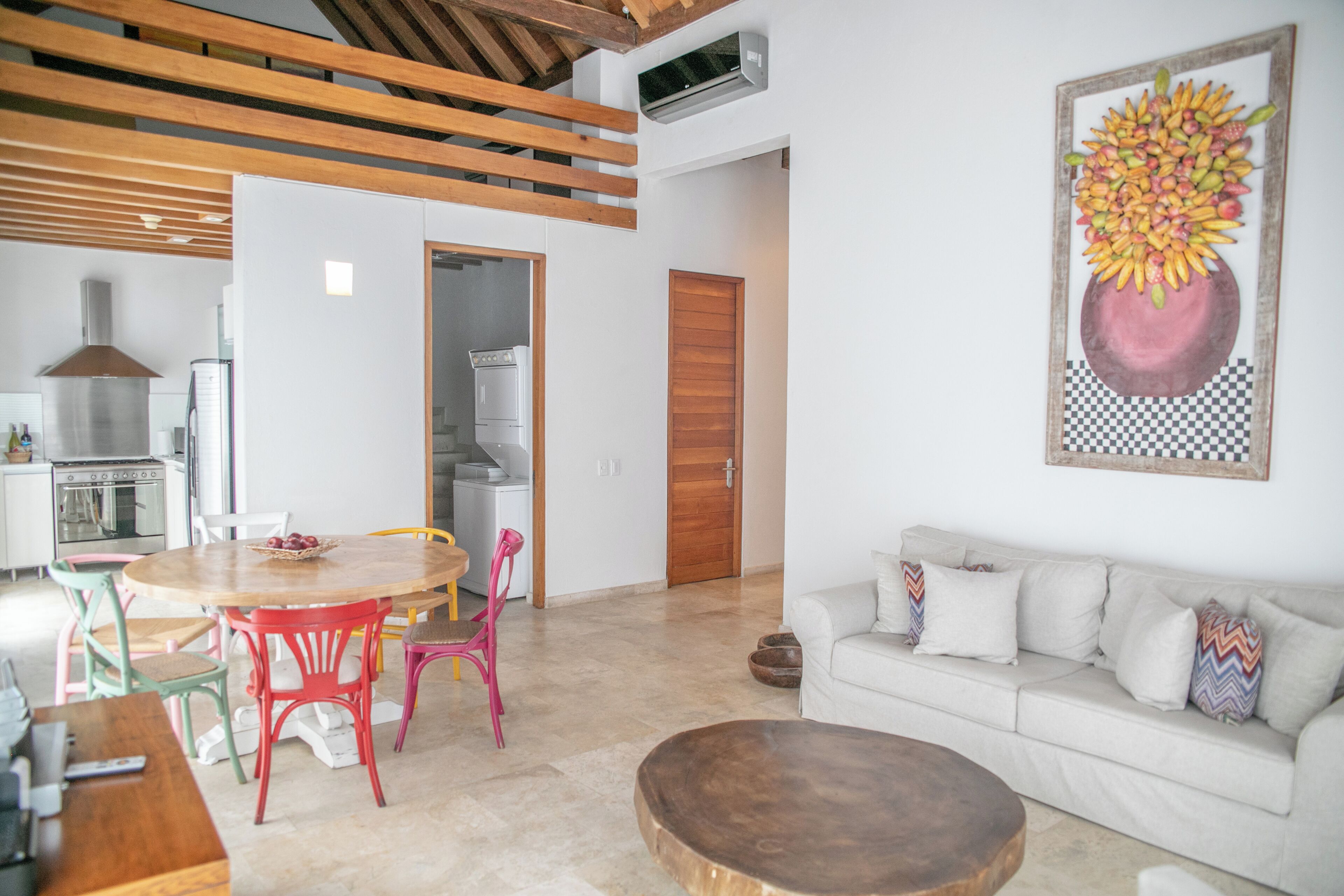 A modern living room with a wooden dining table, colorful chairs, a white sofa, and a large painting of a fruit bowl on the wall.