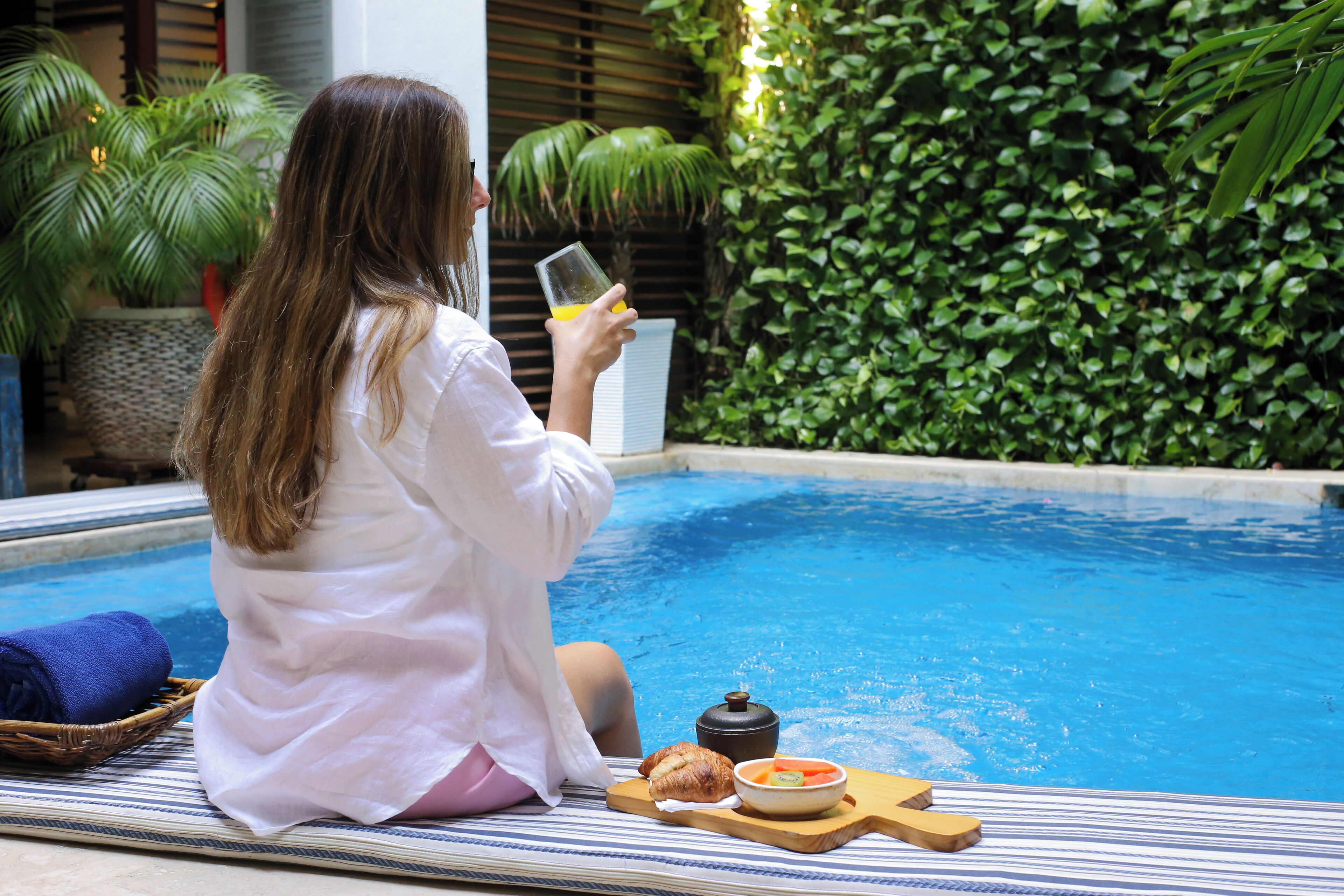 Une personne assise au bord d’une piscine, avec une boisson, un bol de nourriture et une petite casserole sur un plateau.
