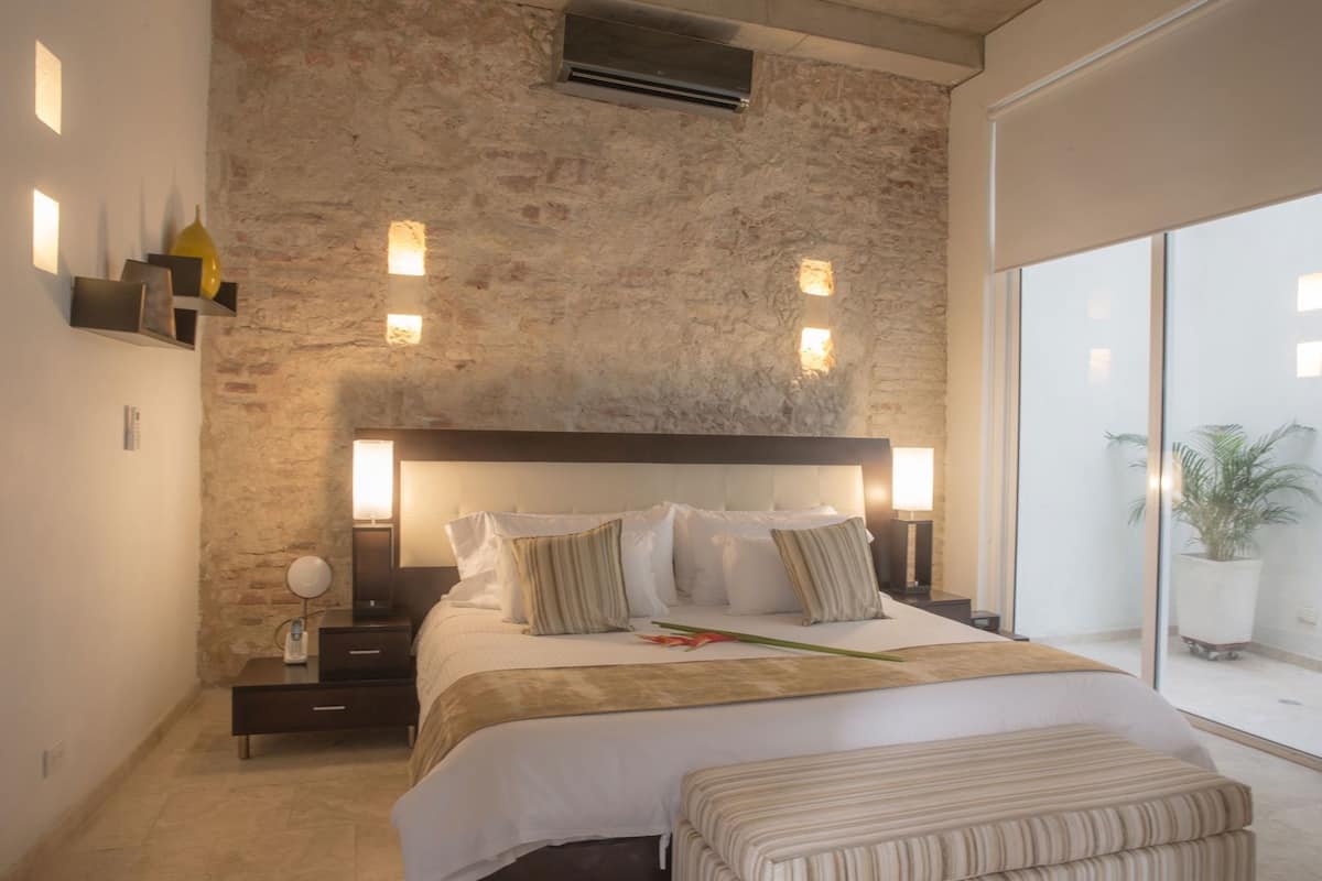 Un dormitorio moderno con pared de piedra, una cama grande con muchas almohadas, mesitas de noche con lámparas y vistas al exterior.