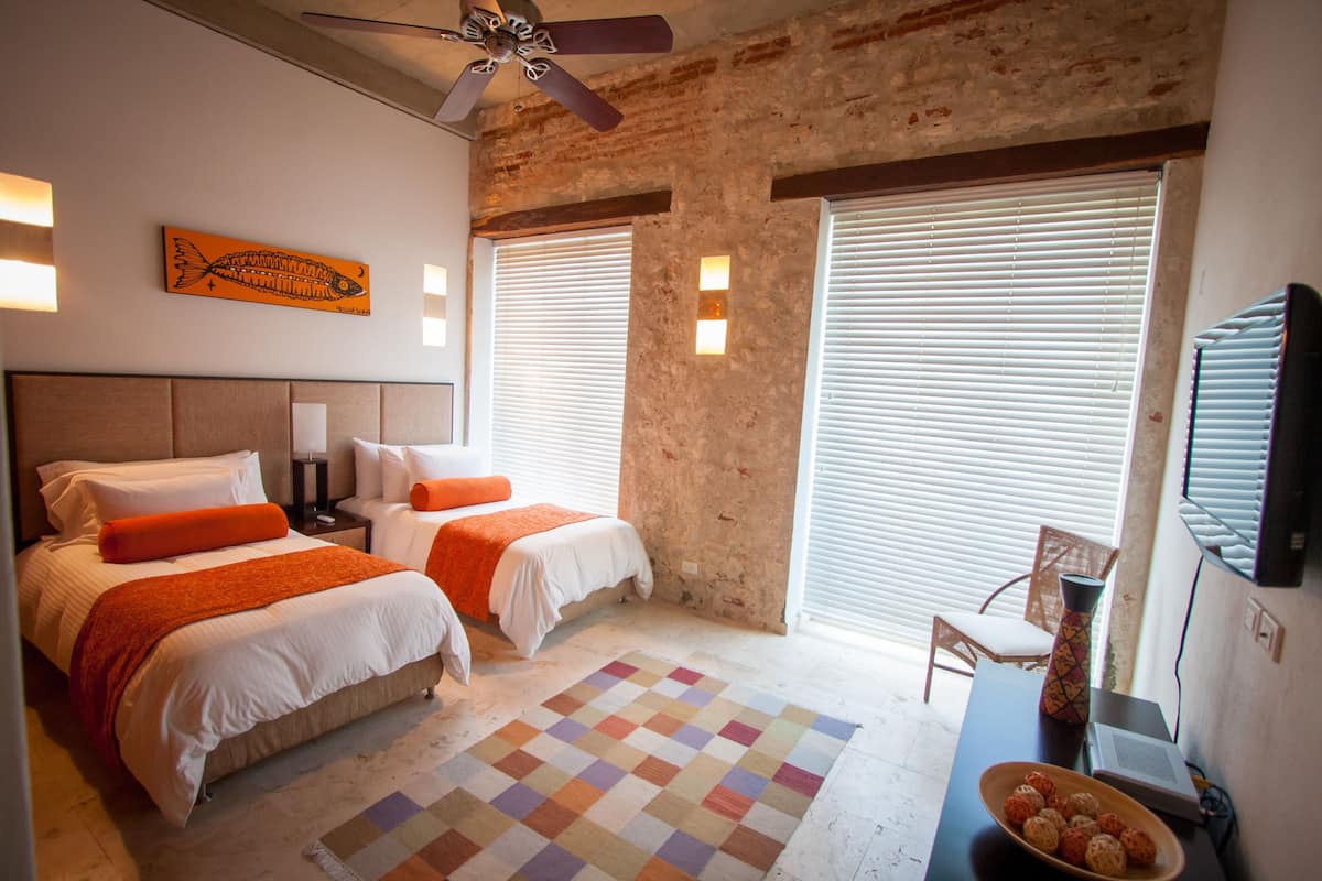 Habitación de hotel con dos camas, un ventilador de techo, alfombra a cuadros y un escritorio con un jarrón y un frutero.