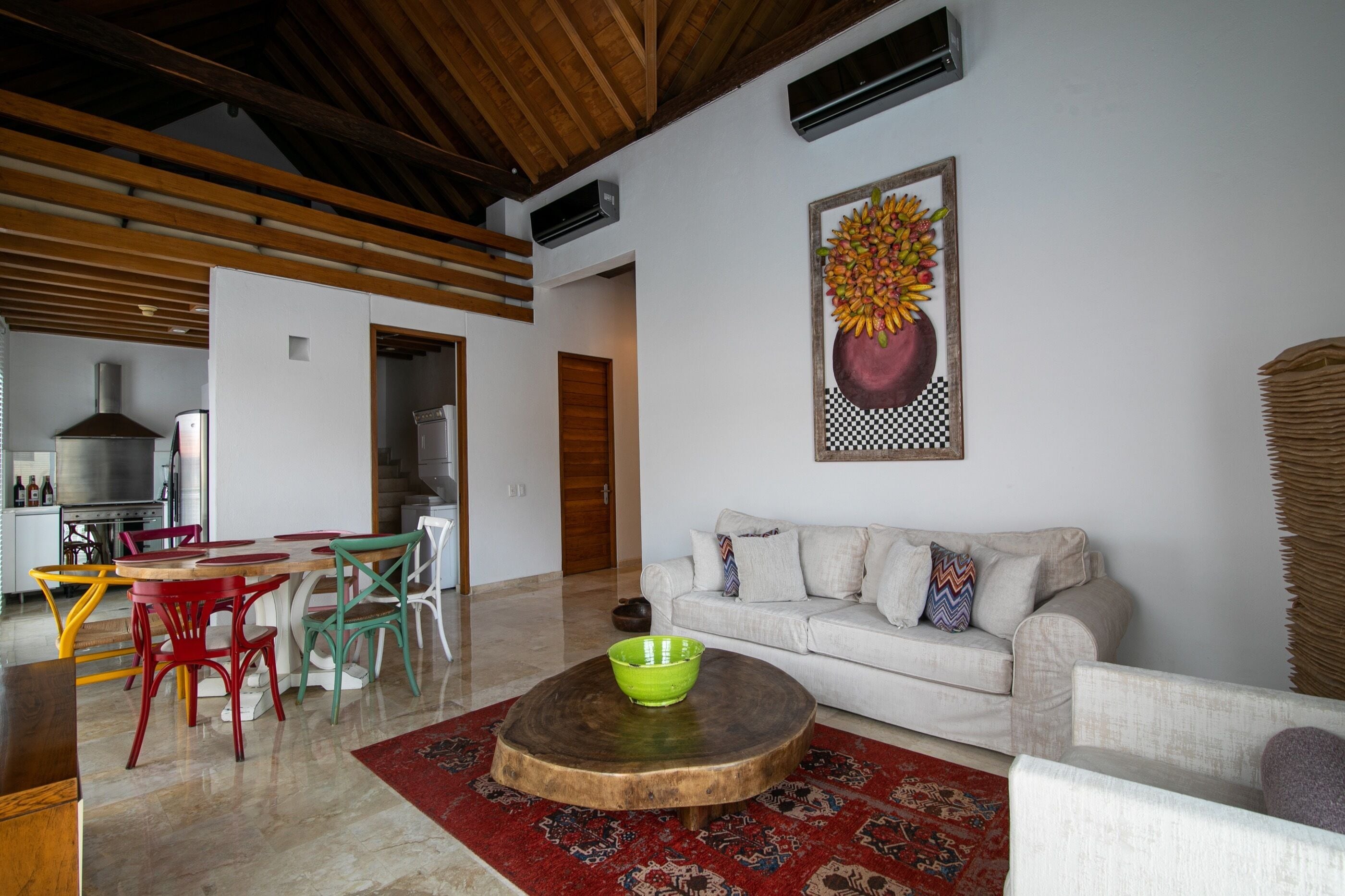 Una sala moderna con techo de madera, comedor con sillas de colores, sofá blanco y un cuadro de un frutero.