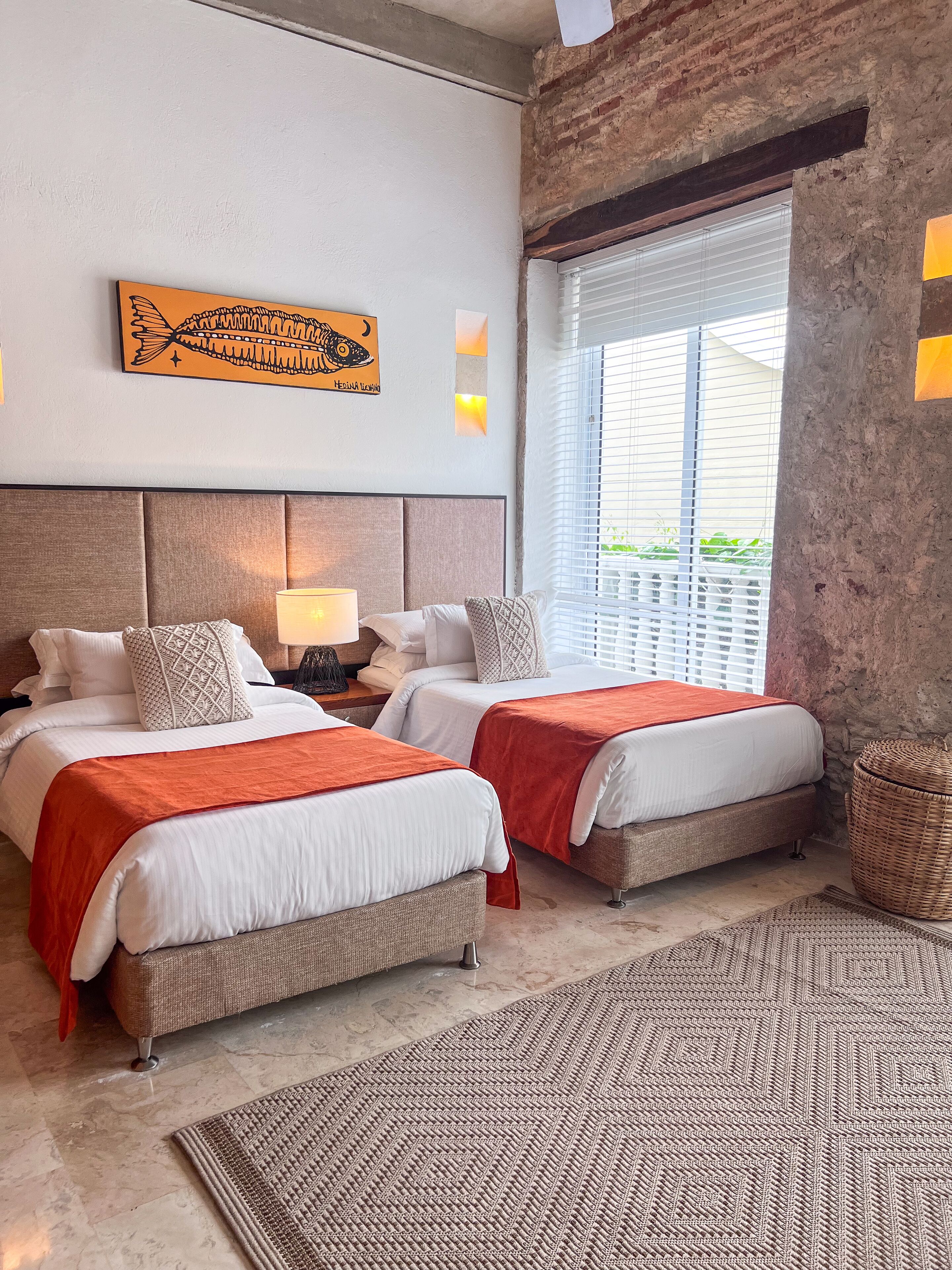 Una habitación de hotel con dos camas, un ventanal con persianas, cabecero, una obra de arte colgada en la pared y una cesta tejida.