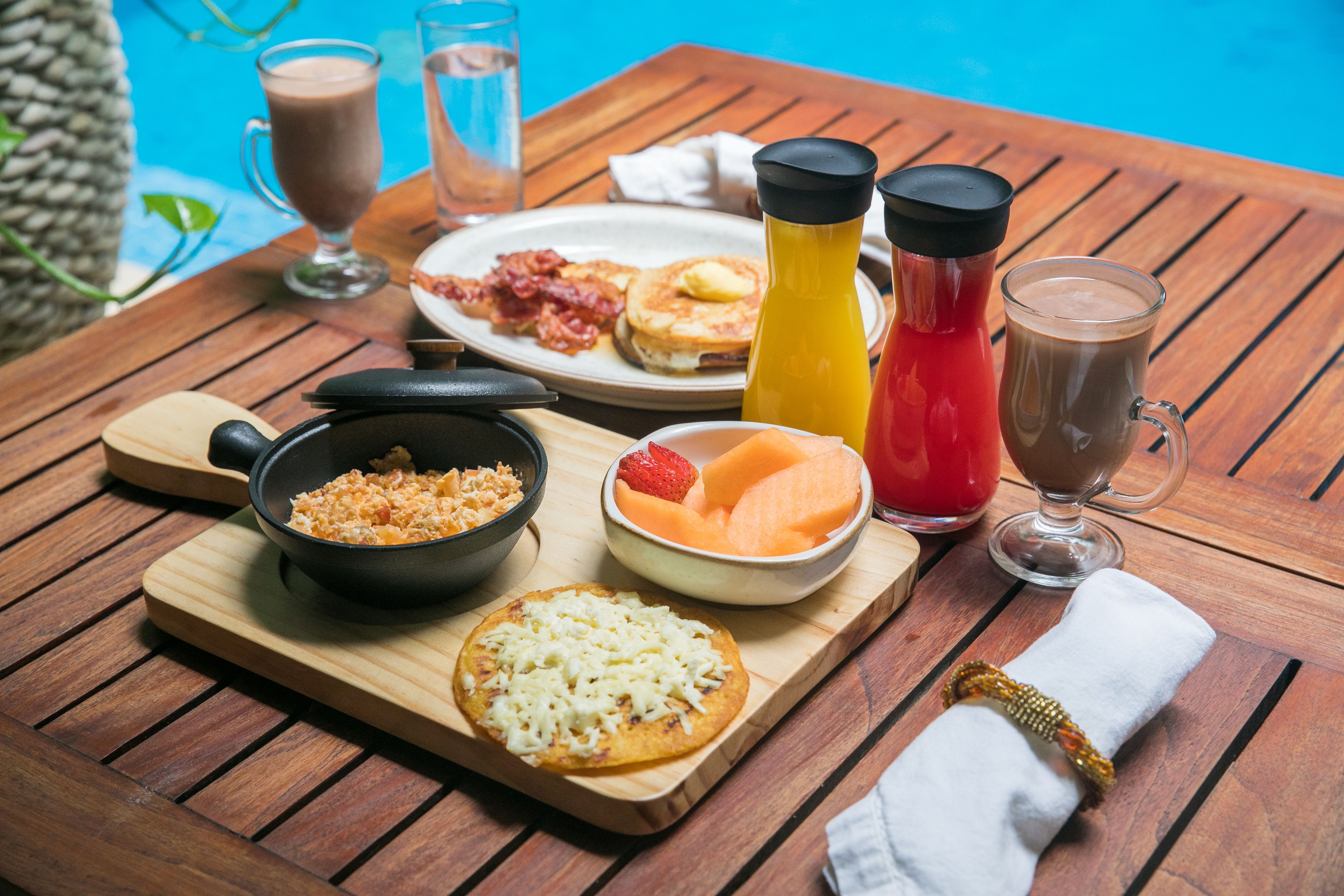 A wooden table with breakfast items including pancakes, a bowl of fruit, a bowl of cereal, and a glass of coffee.