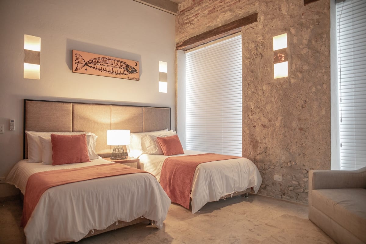 Habitación de hotel con dos camas, cabecero, una obra de arte en la pared, mesita de noche, lámpara y ventana con persianas.