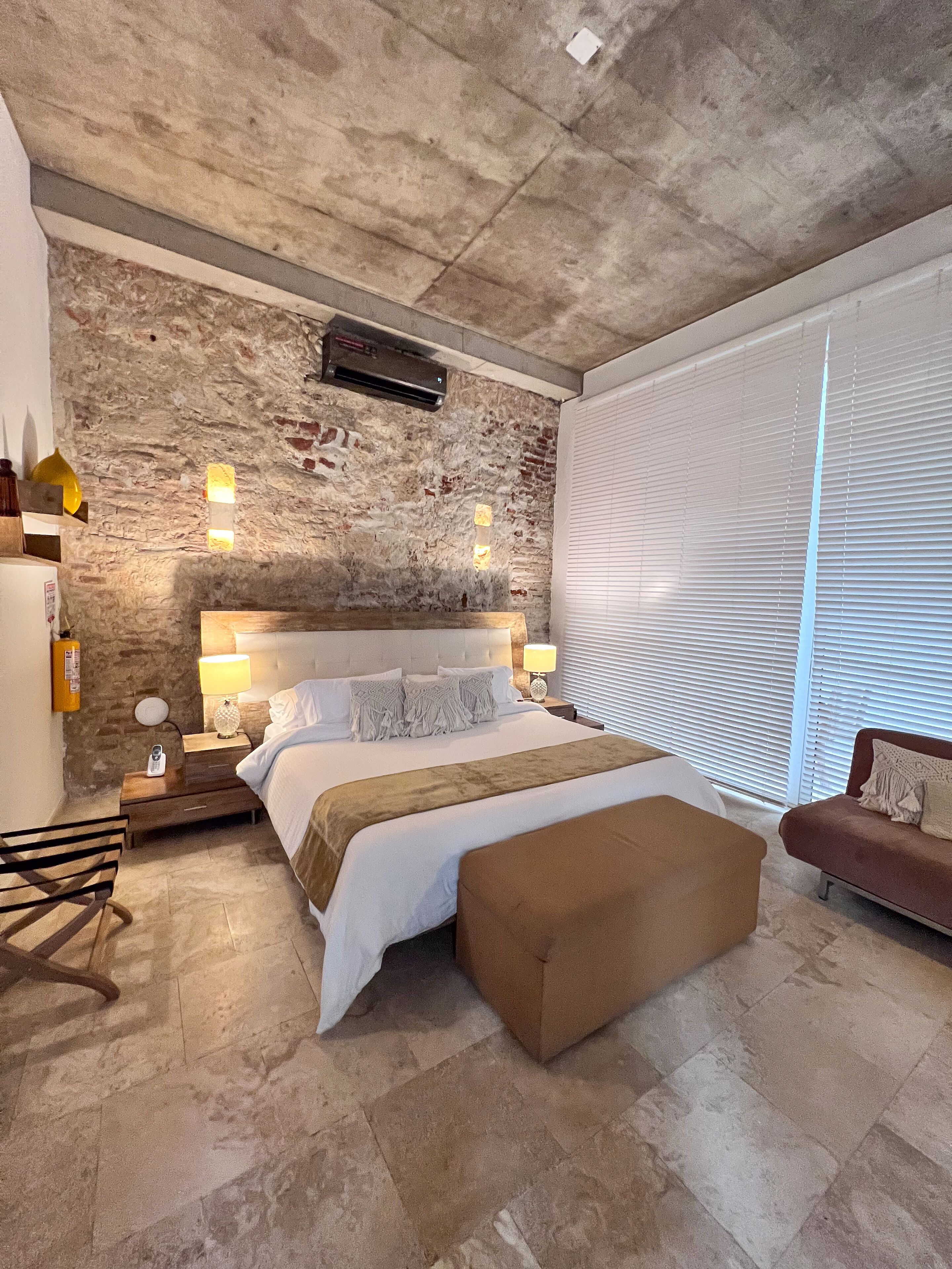 Ein modernes Schlafzimmer mit einem großen Bett, einer hölzernen Bank, einem Nachttisch, einer Lampe und einer Backsteinwand.