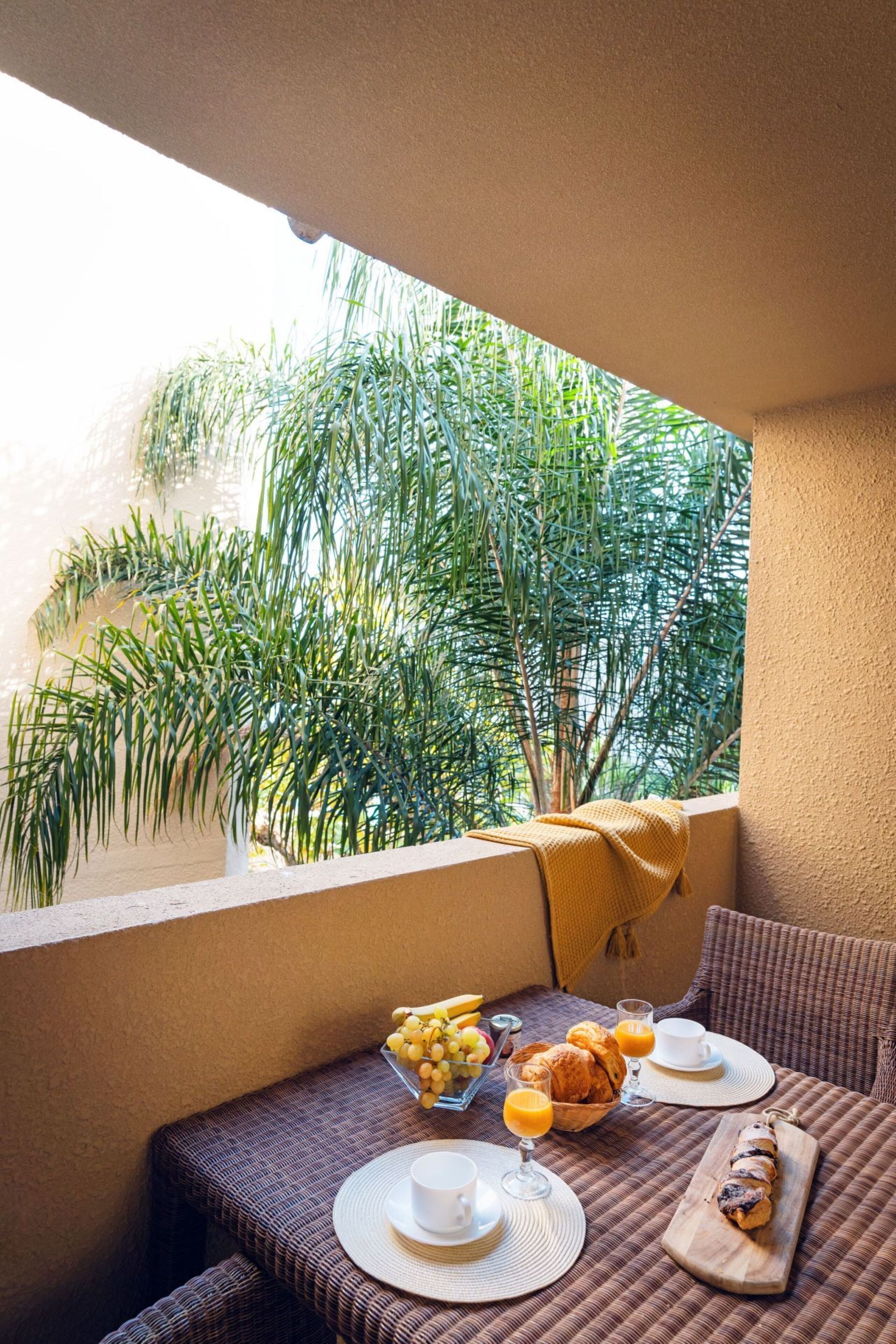A balcony with a wicker table set for breakfast, including croissants, fruit, and orange juice.