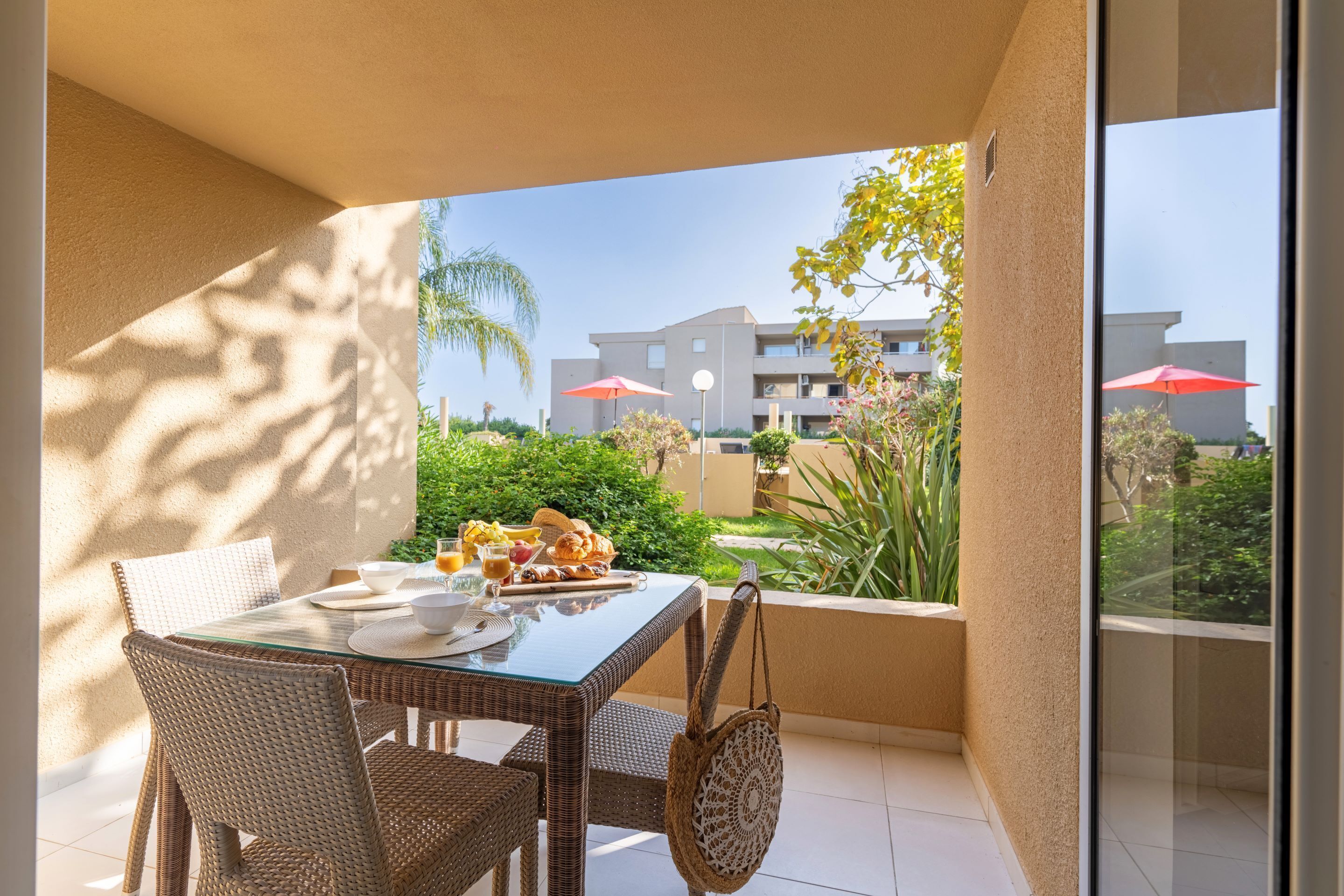 A balcony with a table set for breakfast, wicker chairs, and a view of a building with red umbrellas.
