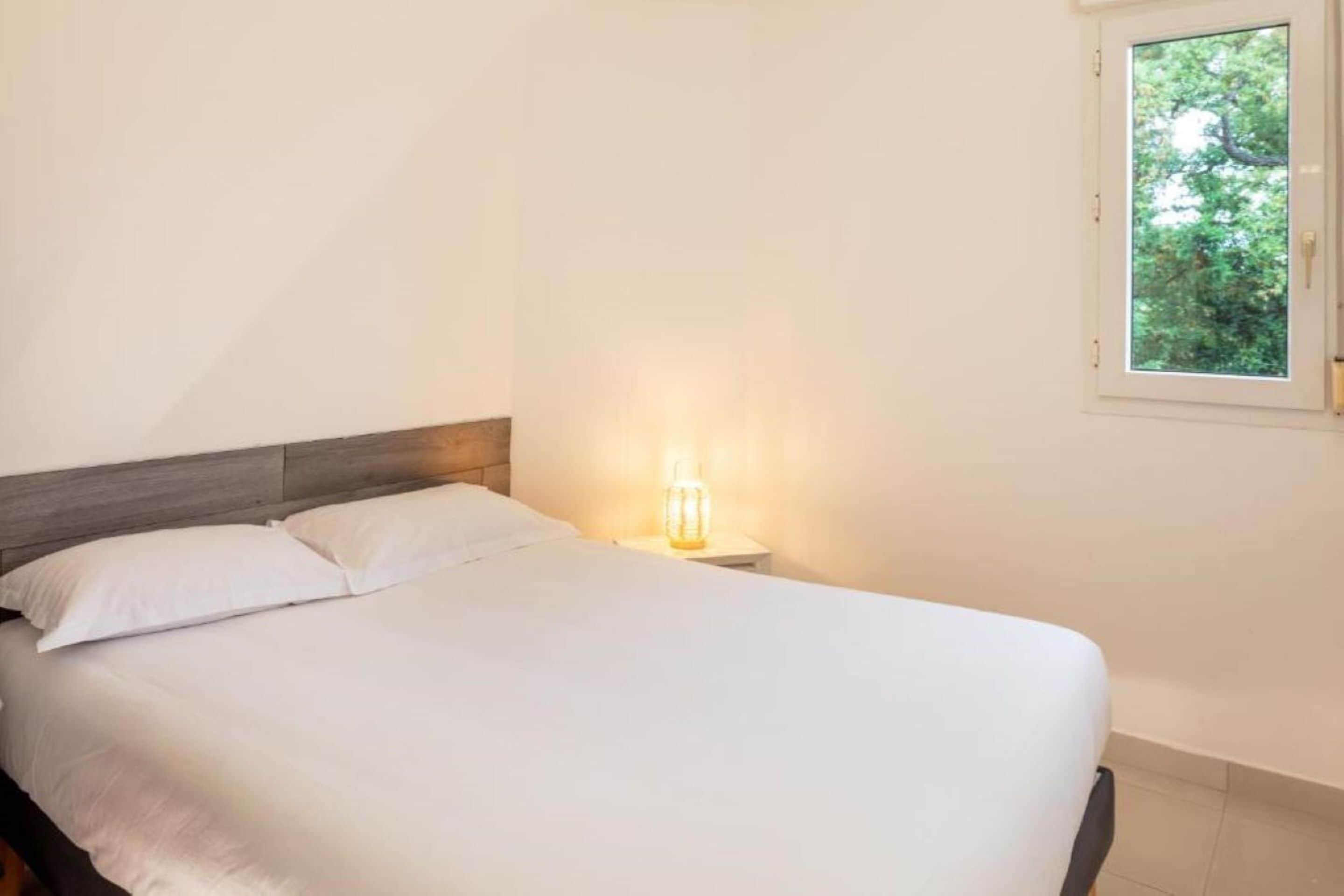 A neatly made bed with white linens in a room with a simple wooden headboard and a small bedside table with a lamp.