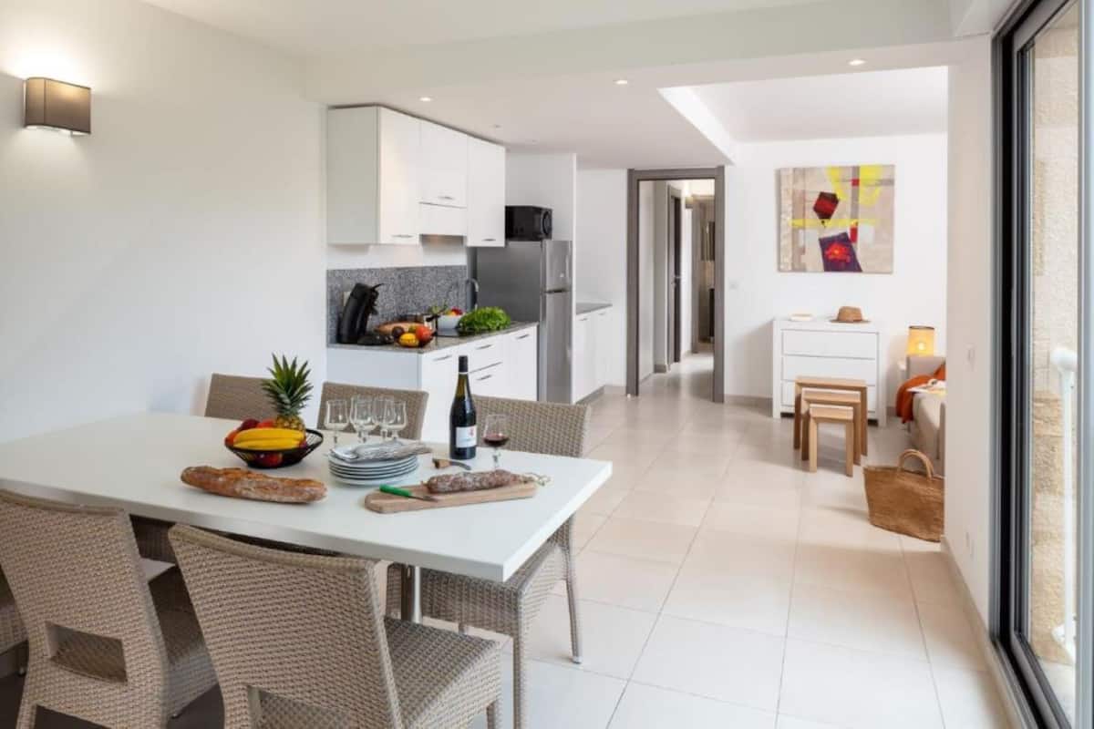A modern kitchen with white cabinets, a dining area with wicker chairs, and a view through a large window.