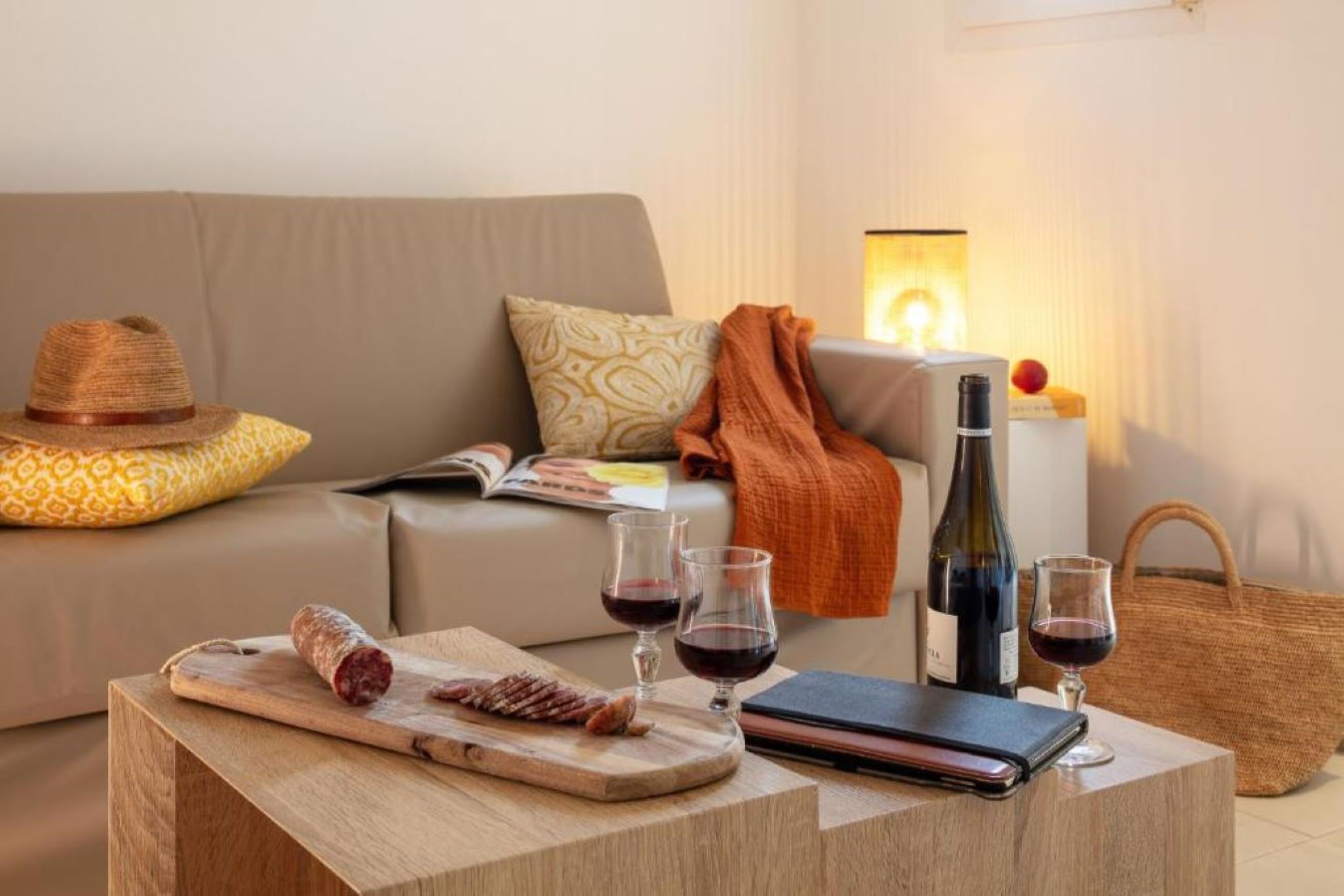 A living room with a sofa, a wooden coffee table with a bottle of wine, glasses, a hat, and a woven bag.