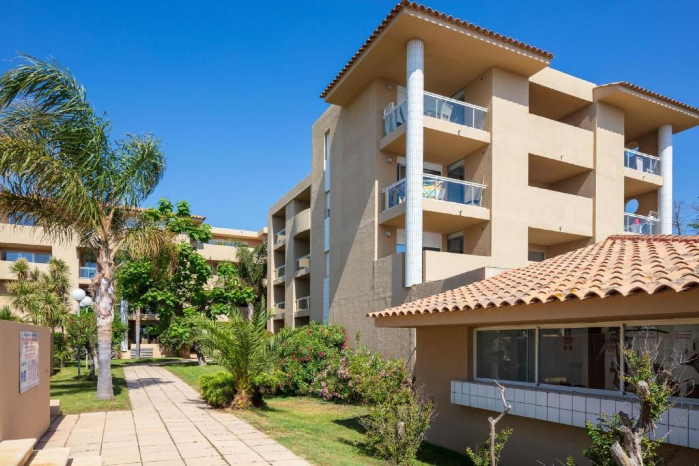 Een hotel met meerdere verdiepingen, voorzien van balkons, een betegeld dak en een verzorgde tuin met palmbomen.