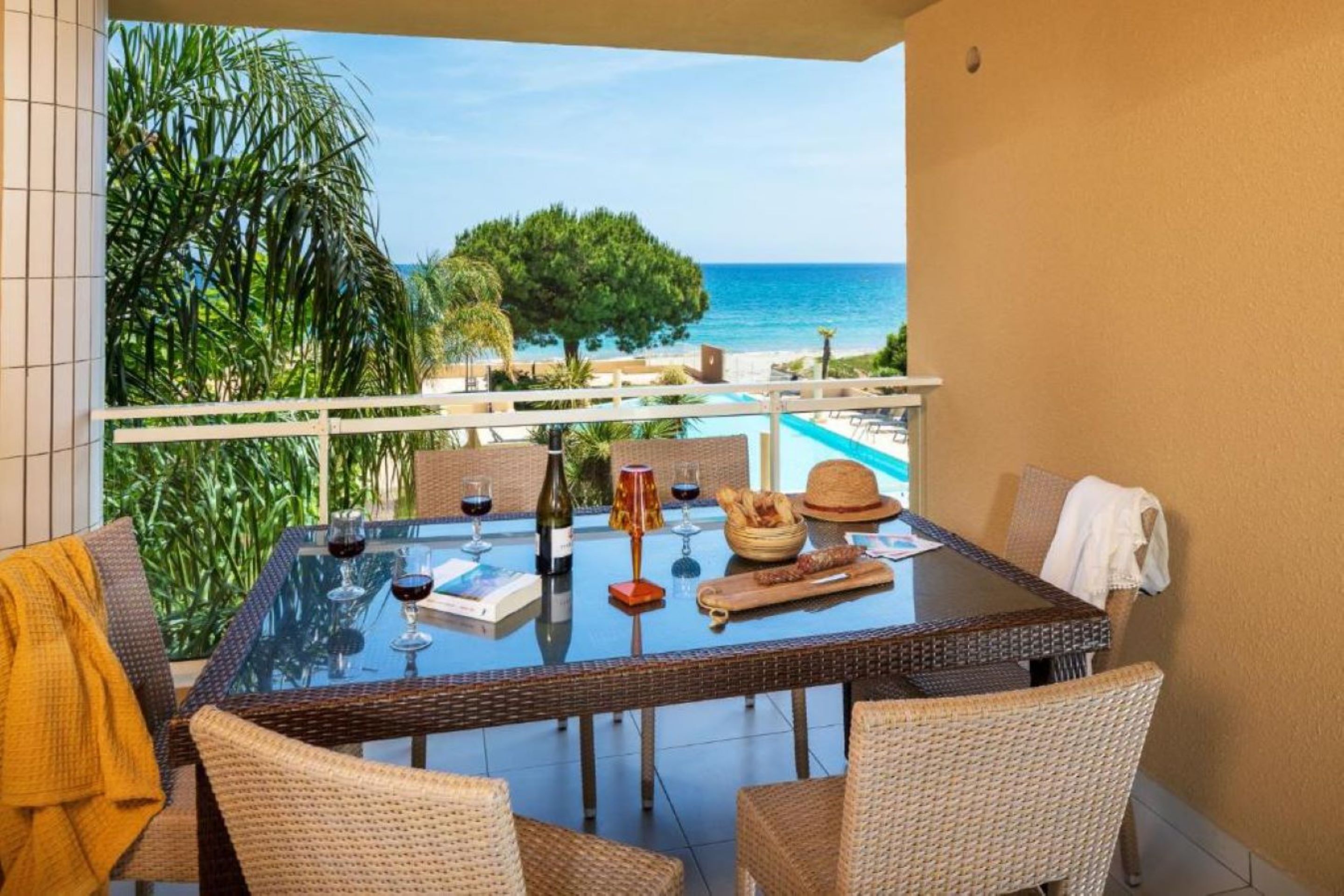Un balcone con un tavolo in vetro apparecchiato per consumare pasti, sedie in vimini e vista sul mare.