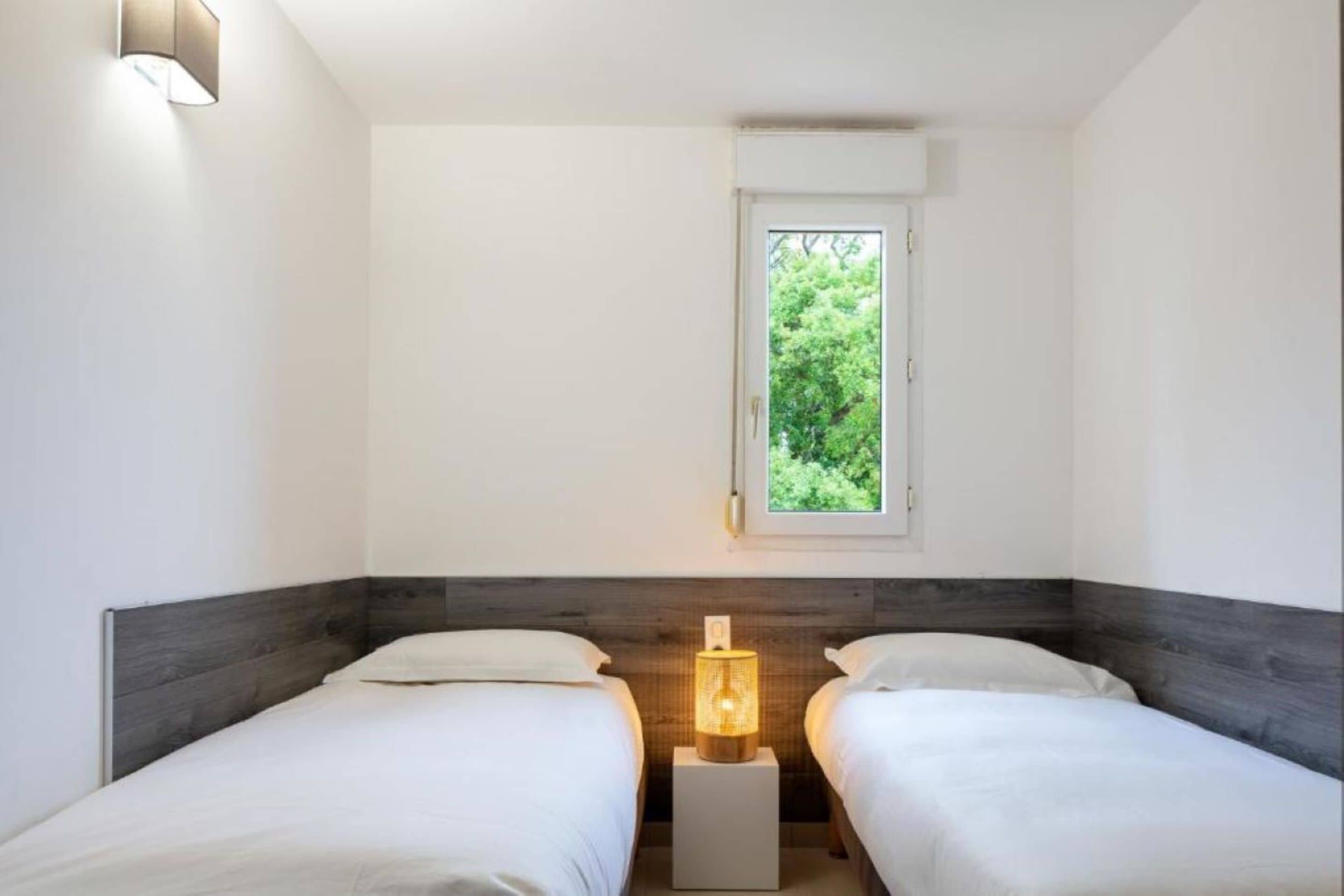 A bedroom with two beds, a window with a view of greenery, and a bedside lamp.