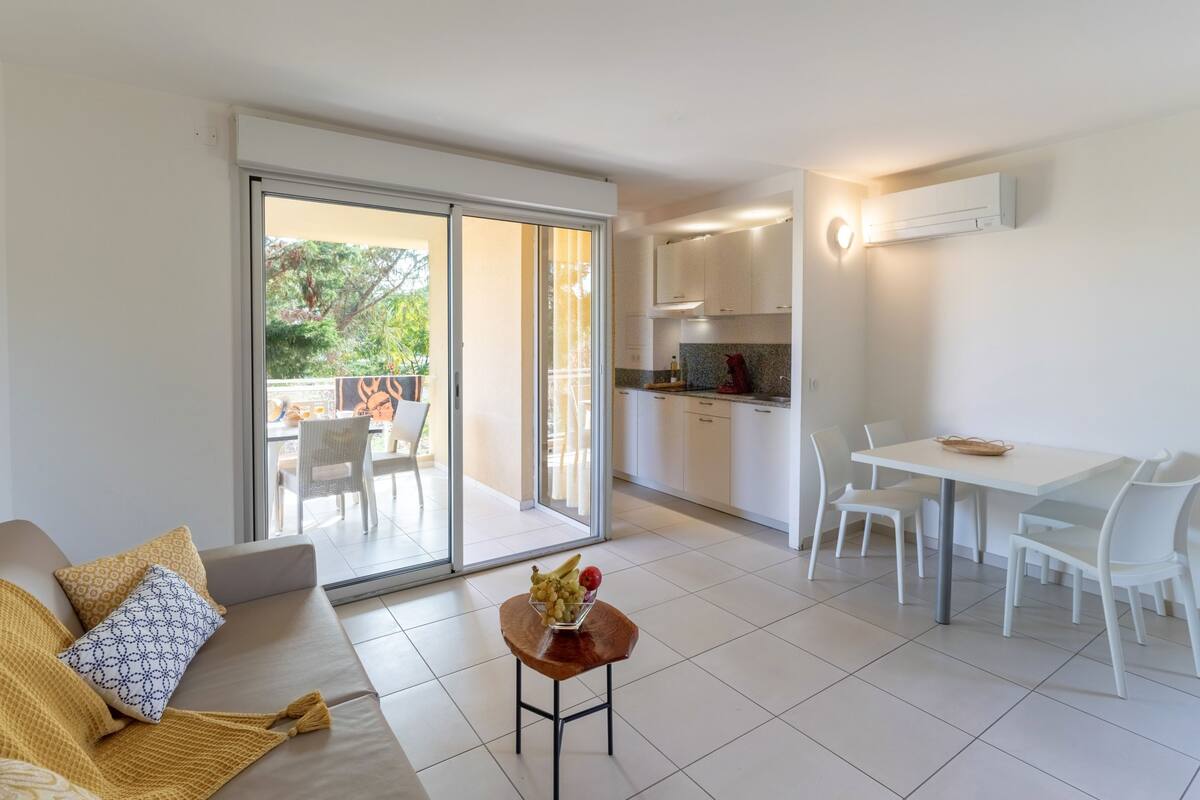 A modern living room with a sofa, a small wooden table with fruit, a dining area with white chairs, and a kitchen with white cabinets and an air conditioning unit.