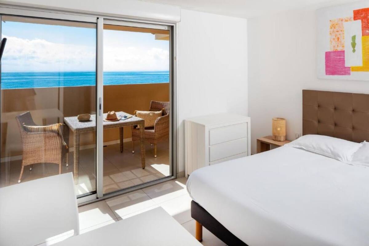 A hotel room with a bed, a desk, and a chair, a sliding glass door leading to a balcony with a view of the ocean.
