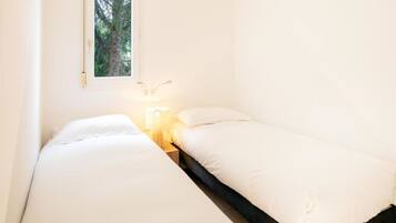 A small, single-bed room with a window overlooking a tree, a bedside table with a lamp, and a wall-mounted light fixture.