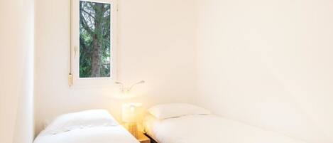 Una habitación pequeña con cama individual, ventana con vista a un árbol, mesita de noche con lámpara y un aplique de pared.