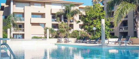 Une piscine avec des chaises longues et un bâtiment doté de balcons, avec des palmiers en arrière-plan.