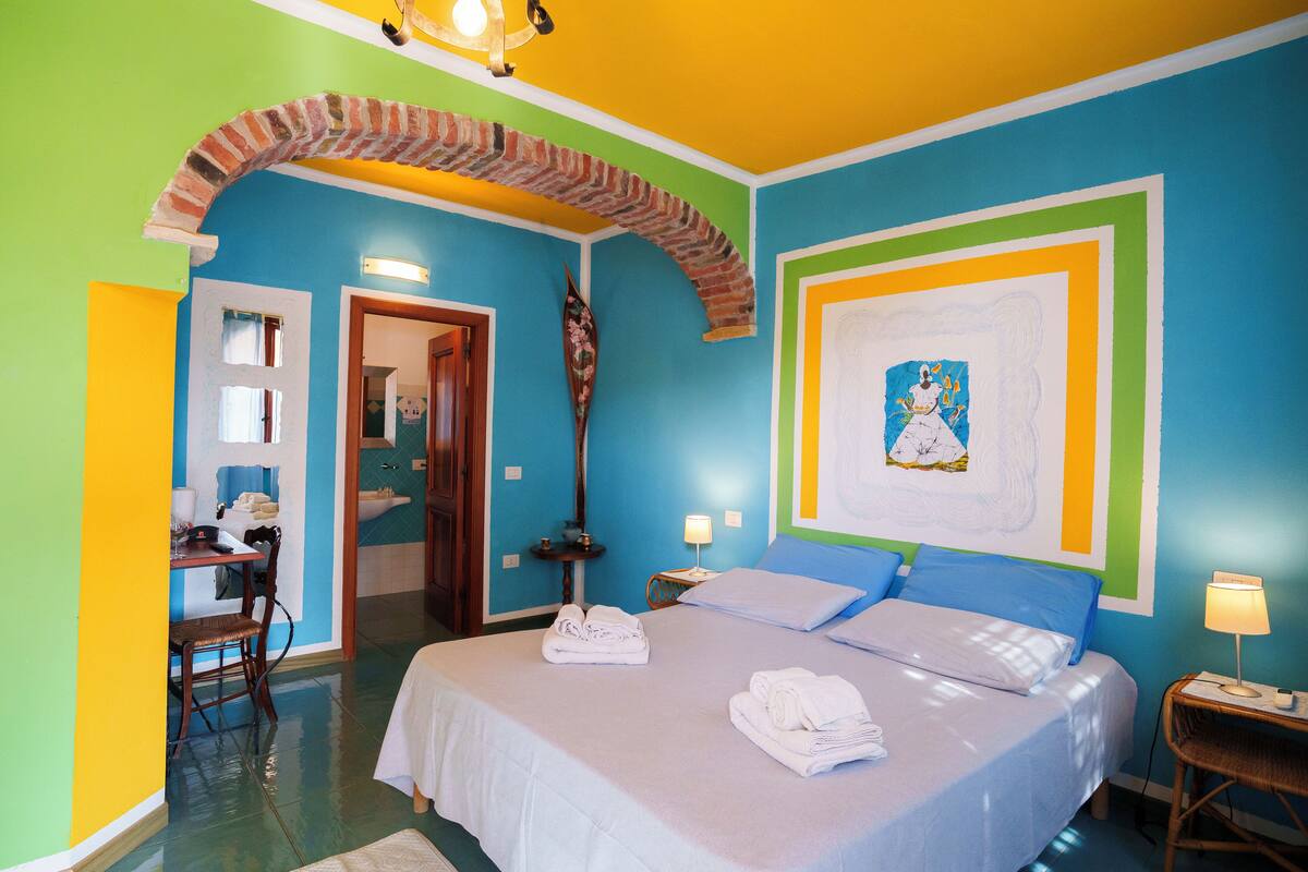 A room with a bed, a desk, and a chair. There is a door leading to another room.