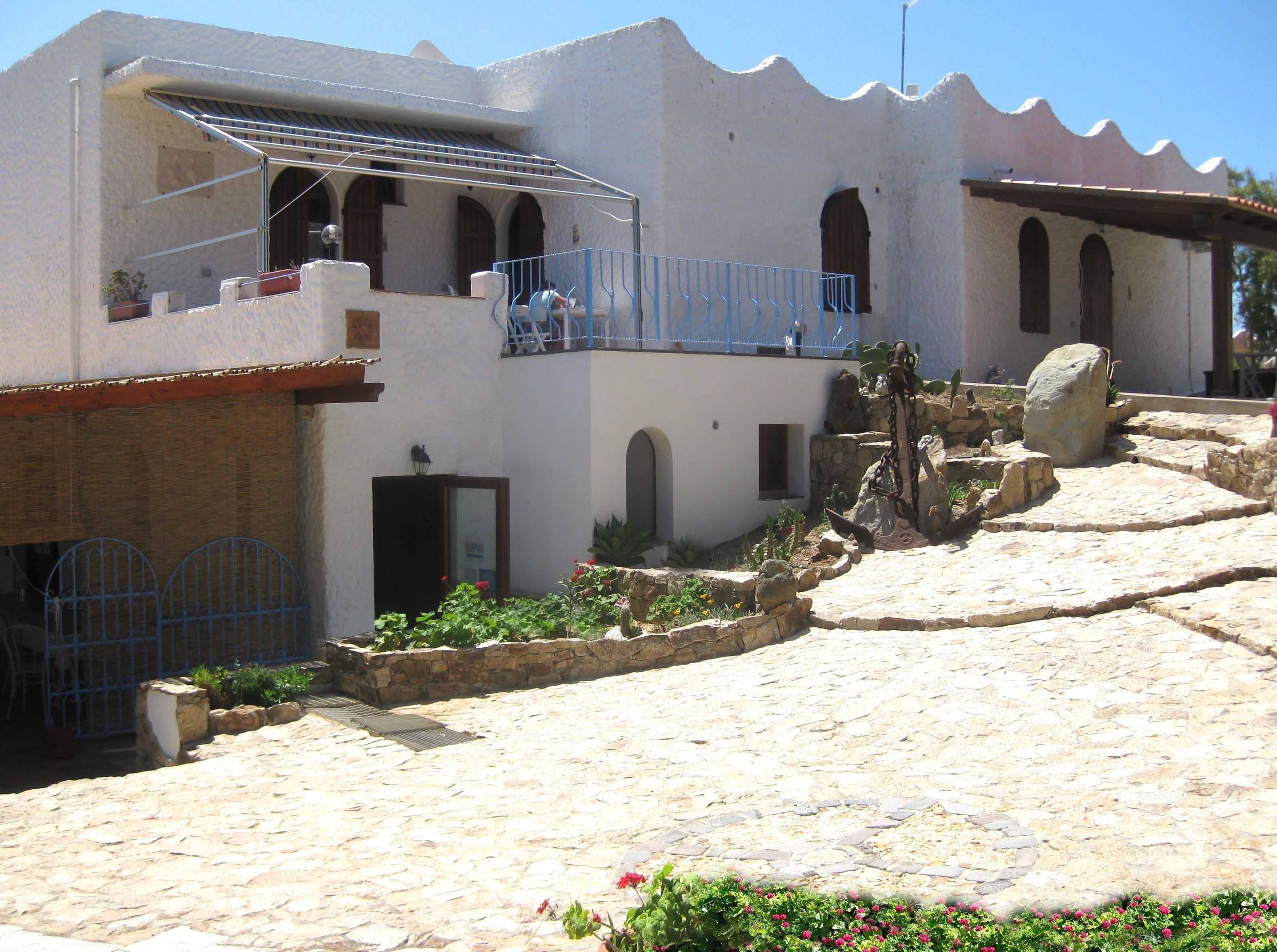 Un bâtiment de style méditerranéen traditionnel, doté d’une cour en pierre, de portes en arc de cercle et d’un balcon avec des balustrades bleues.
