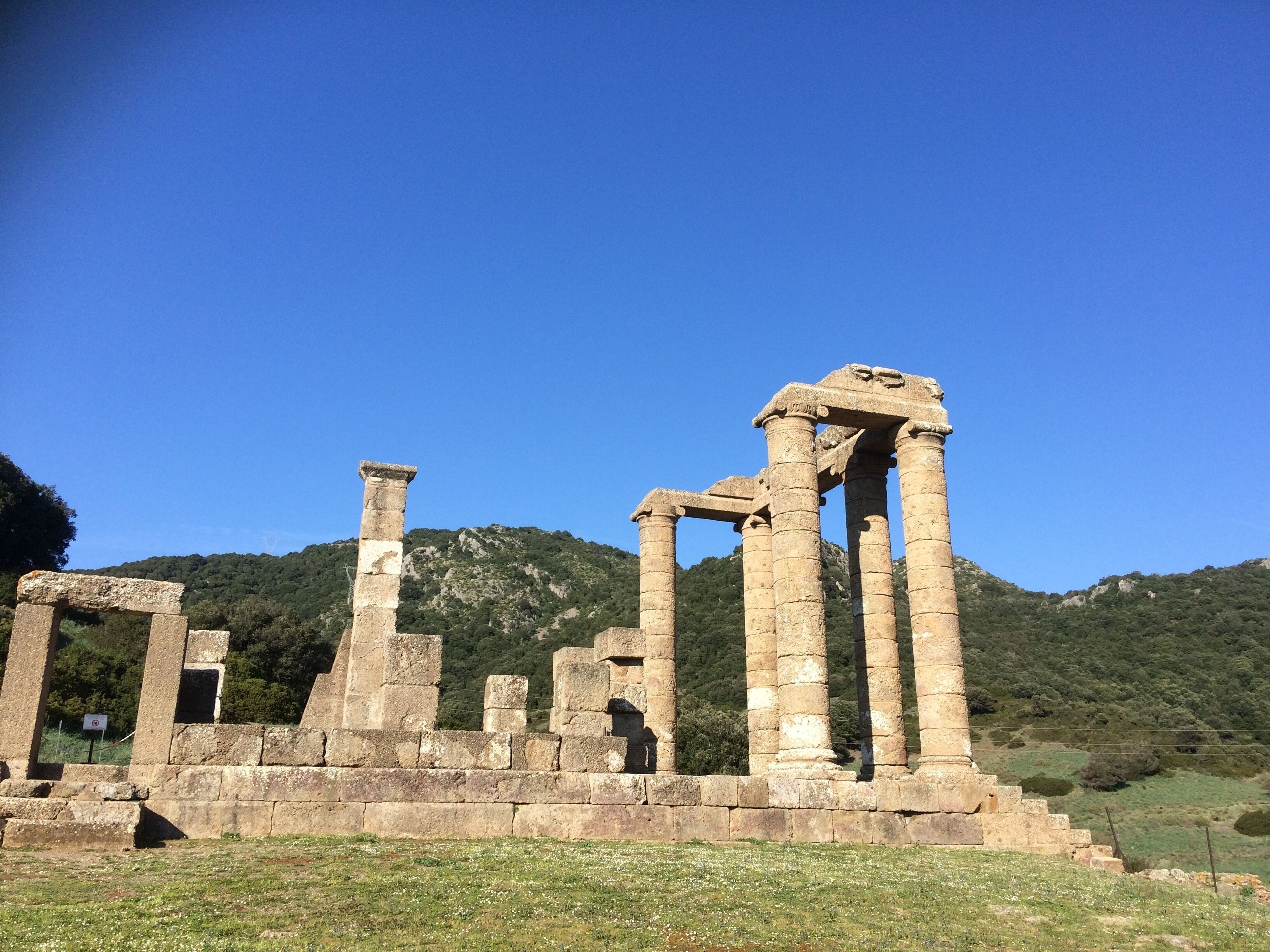 Ruinas de piedra antiguas con columnas y una estructura pétrea, frente a colinas verdes y un cielo azul despejado.