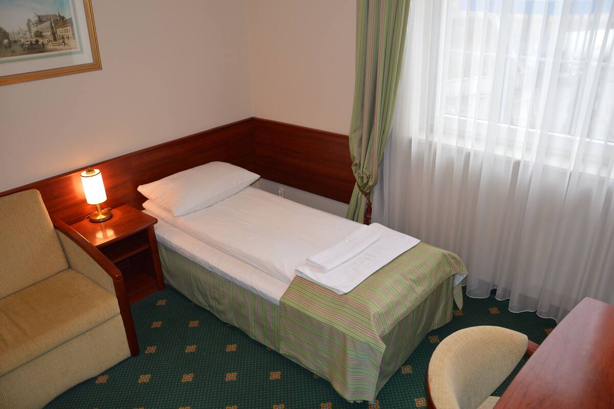 Una habitación de hotel pequeña con una cama individual, una mesita de noche con lámpara, una silla y una mesa.
