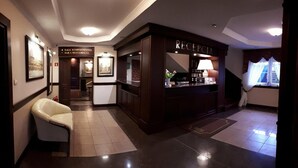 Un vestíbulo de hotel con una recepción, obras de arte enmarcadas en las paredes, una butaca de cuero blanco y un cartel que indica la sala de conferencias y el bar.